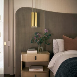 Portfolio: A rounded wood nightstand is styled with fresh florals next to a sculpted headboard and beneath suspended brass pendants.