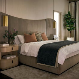 Portfolio: A sweeping upholstered headboard anchors the primary bedroom, complemented by brass pendant lighting, rounded nightstands, and softly layered textiles