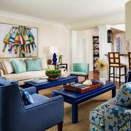 A neutral room is brightened by paired blue coffee tables & matching armchairs.