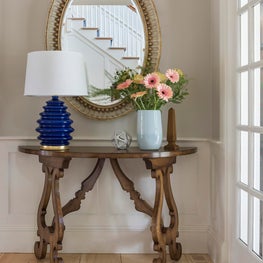 Demilune carved table, blue lamp, oval mirror, striped rug entryway