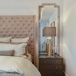Blush upholstered bed with framed floor length mirror and chic bedside lamp.