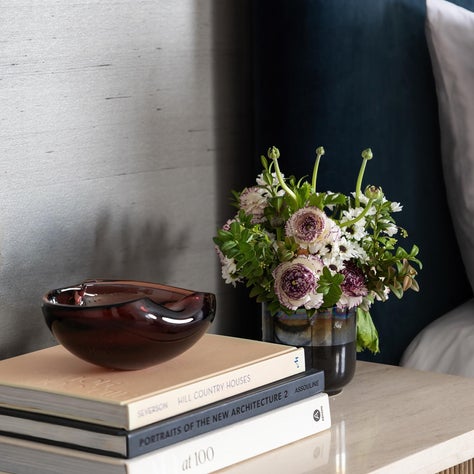 Laura U Design Collective, cover: A vignette of the bedroom pairs a reeded wood nightstand with a stone top, layered books, fresh florals, and warm brass accents.