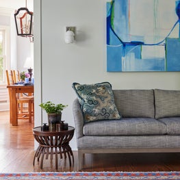 Kenilworth Living Room. Blue textured sofa with vibrant artwork, modern sconce, oriental rug & sculpted wood side table.