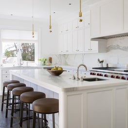 White kitchen with leather bar stools, brass pendants, and woven window shades.