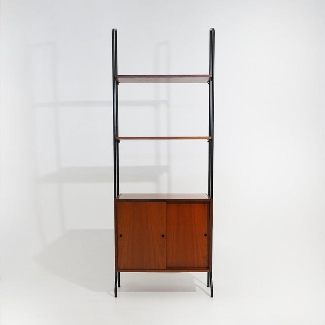 The lightness of the black lacquered metal frame and the warmth of teak define this modular bookcase produced by Amma...