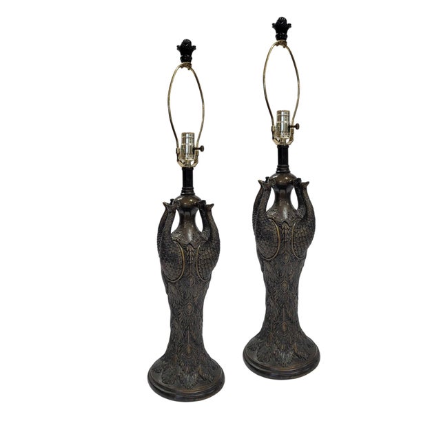 A pair of sculptural table lamps centered on an elegant peacock motif. Each lamp features a classical urn-shaped body...