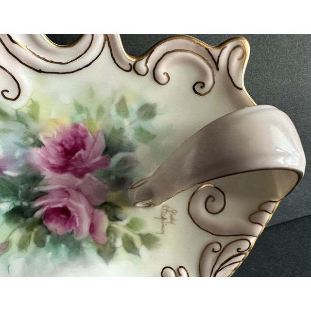 Pretty trinket dish that is shell shaped with a handle and hand-painted in a floral motif of pink roses. Gold gilding with...
