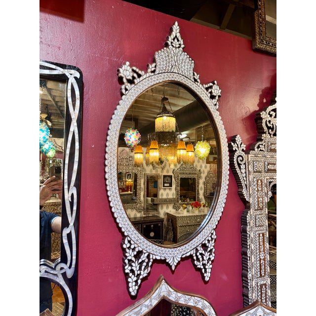 All white mother-of-pearl inlaid wall mirror featuring an elegant oval silhouette and ornate crown crest. The frame is...