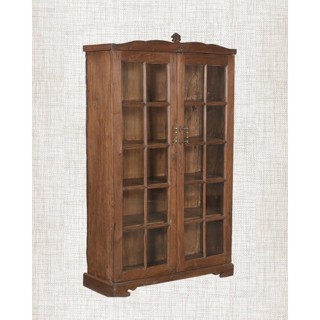 A carved finial at the crown. An arched pediment. Ten individual glass panes per door. This antique Indian armoire is...