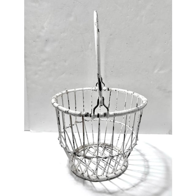 Vintage wire egg gathering basket with a tall swing handle, tapered sides and a worn white painted finish. The open wire...
