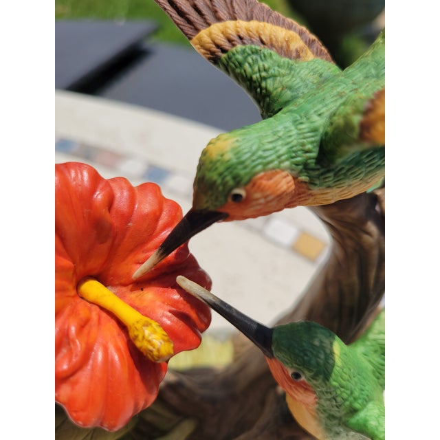 A beautifully detailed porcelain hummingbird sculpture, featuring two vibrantly painted birds hovering around a red...