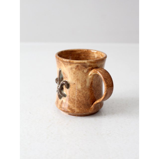 A vintage studio pottery mug featuring a raised fleur-de-lis motif on one side. The glaze blends golden brown and cream...
