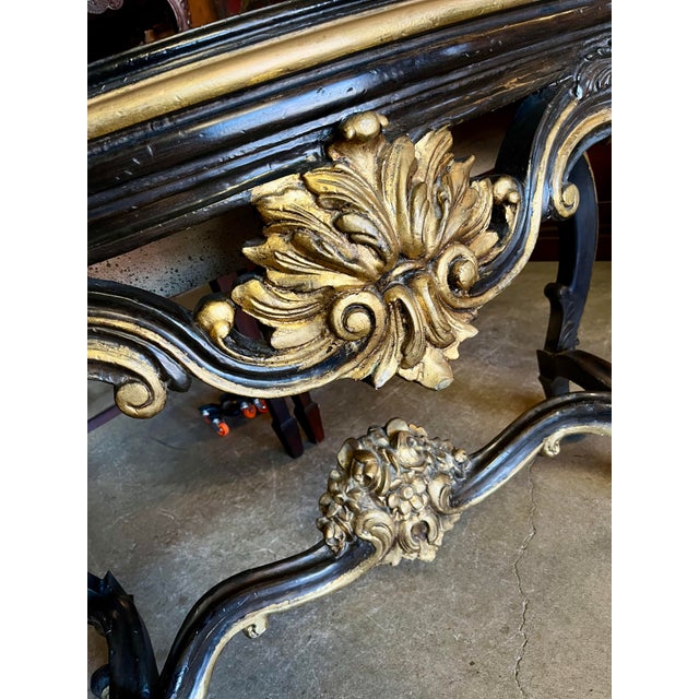 This baroque-style console table features an ornately carved black wooden frame with gilded accents, exuding Old World...