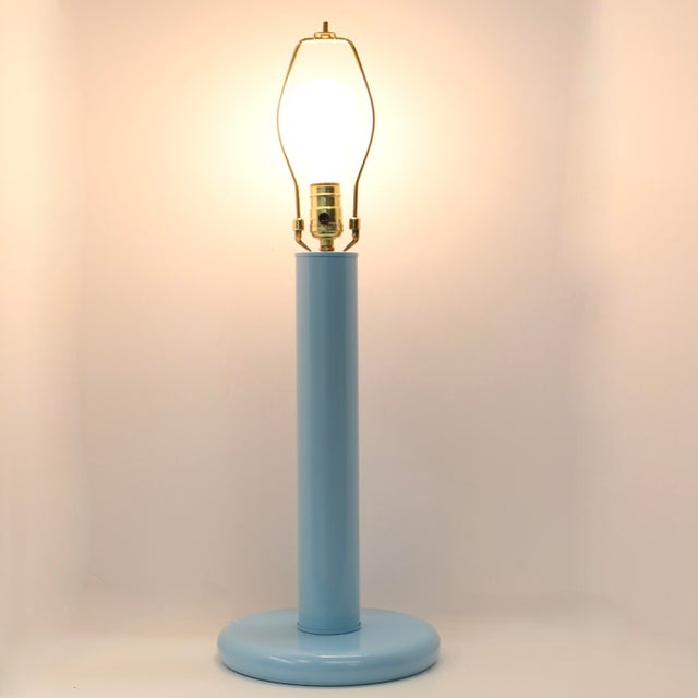 The lamp features a slender, tubular neck transitioning into a weighted circular "puck" base. Its understated silhouette...