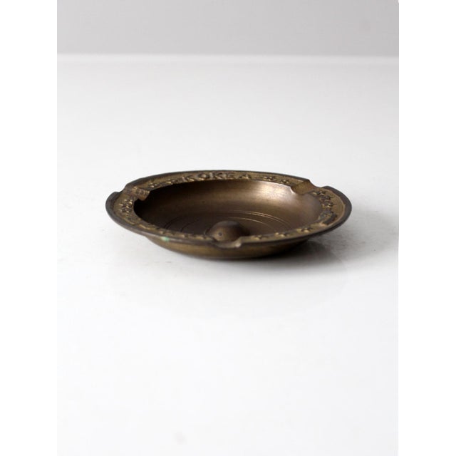 A vintage brass ashtray marked “KOREA” featuring a ring of raised stars along the rim. The low, round form has substantial...