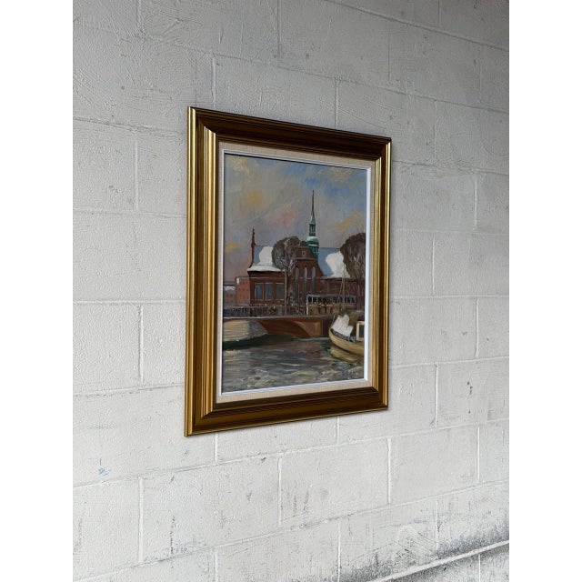 This scenic landscape painting depicts a tranquil winter canal setting with snow-capped architecture and a moored boat....
