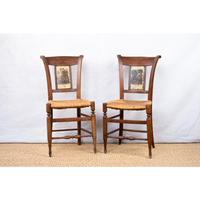 A pair of antique chairs with rush seats and hand painted splats, late 19th century, French. Splats feature two scenes...