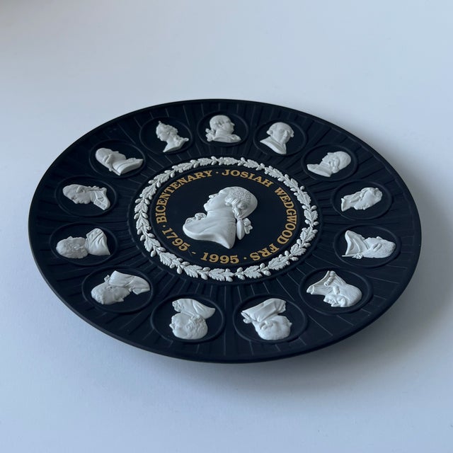 This circular plaque features a matte black stoneware body with applied white bas-relief details. The central medallion...