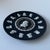 This circular plaque features a matte black stoneware body with applied white bas-relief details. The central medallion...