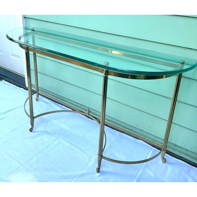A 1960s Hollywood Regency demi-lune console table with a brass-tone metal frame and substantial beveled glass top. Its...