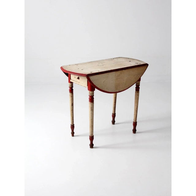 An antique drop leaf table. The end table features turned legs and front drawer. The leaves have wood holds. Original red...