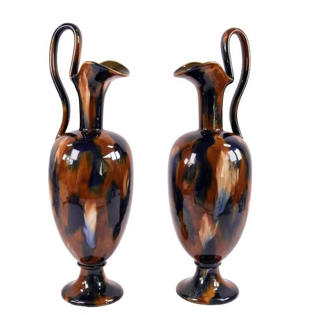 High looping handles sweep prominently above the pinched pouring lips of this Victorian pair of decorative ewers. Each...