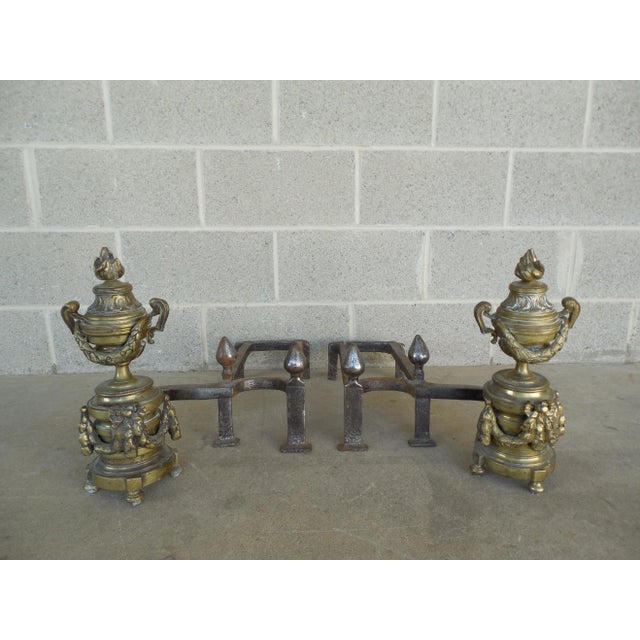 Pair of brass and iron andirons. These feature hand-crafted iron construction and detailed, ornate brass urns with flame...