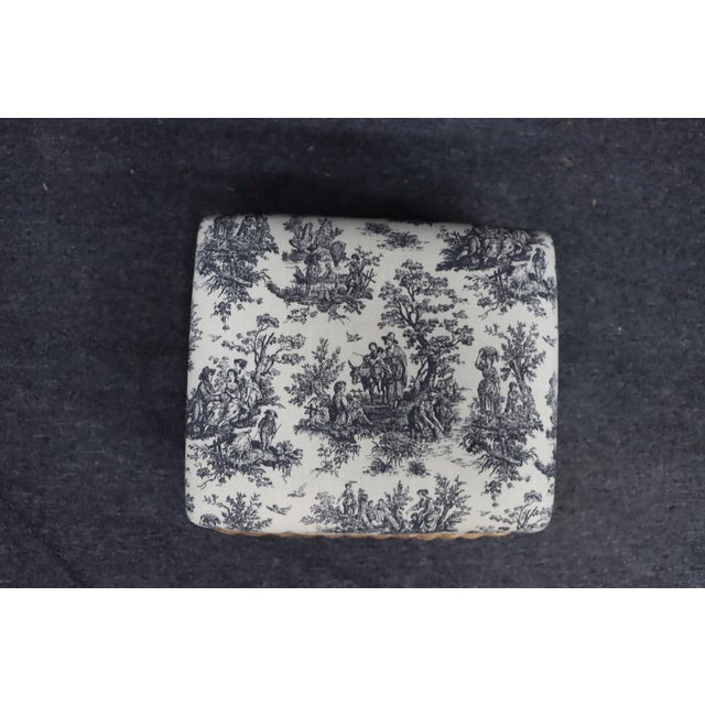 Wicker ottoman with a woven base and upholstered cushion. Features a black and cream toile fabric with a classic scenic...