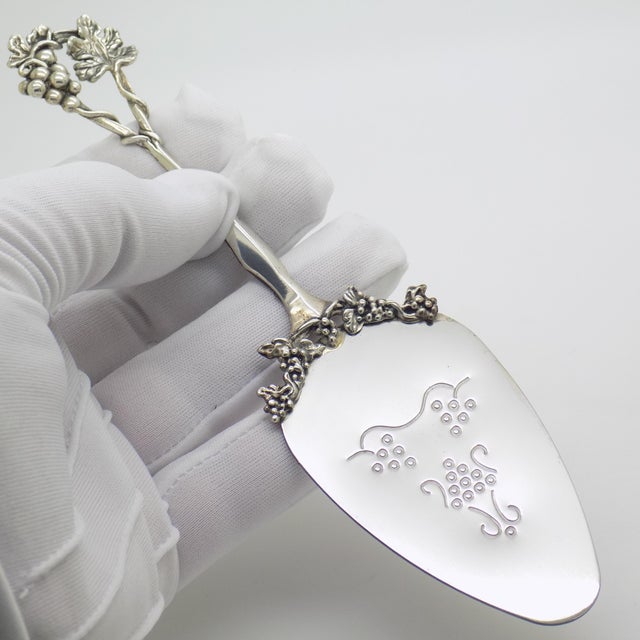 This vintage silver tableware object depicts a cake server with a grapevine and grape cluster relief handle, crafted as a...