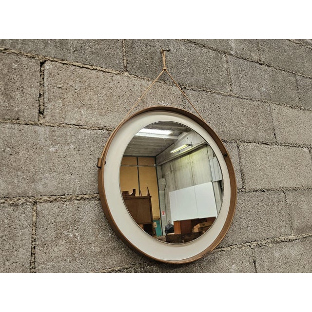 1970s wall mirror with a brown wood-effect PVC frame and faux leather interior, held by a cord for wall hanging. Normal...