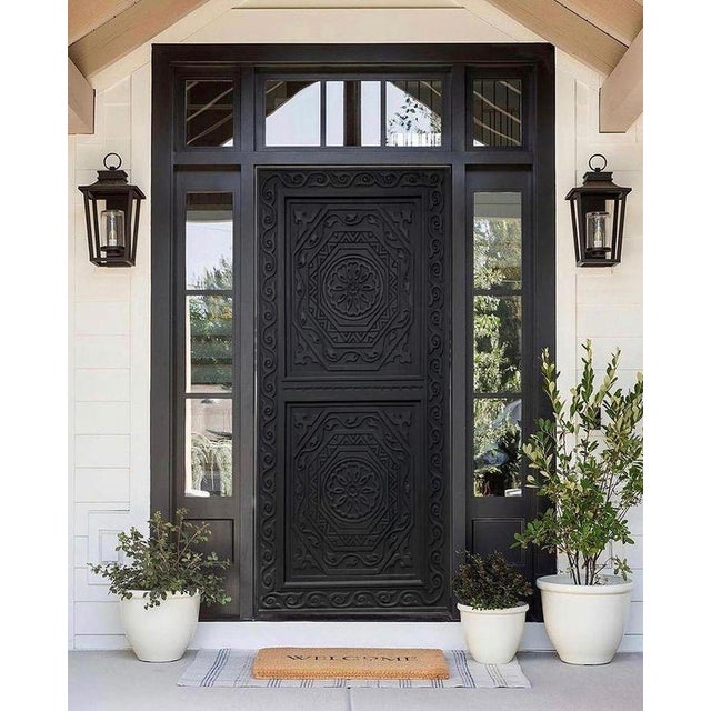 This handcrafted black solid wood entry door features deeply carved geometric and floral motifs with a symmetrical layered...