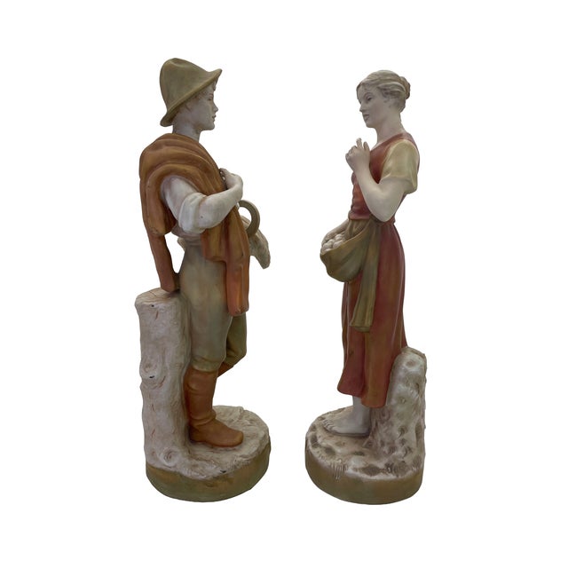 A pair of Royal Dux porcelain harvest figures depicting a young man carrying a sheaf of wheat and a sickle, with a cloak...