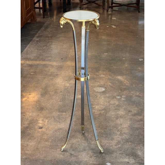 Three curved steel legs headed by brass rams' heads and ending in brass hoof feet, joined by a brass ring, support a small...