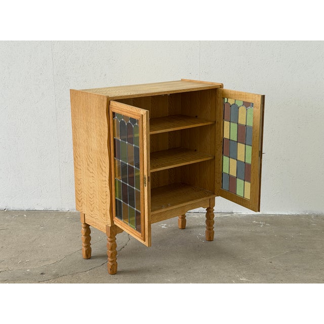 A captivating solid oak display cabinet with leaded stained glass doors in the style of Henning Kjærnulf, combining the...