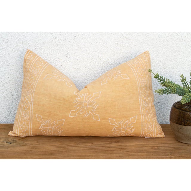 A four-petal flower-star medallion, hand block-printed in white, repeats across a warm golden linen ground on this lumbar...