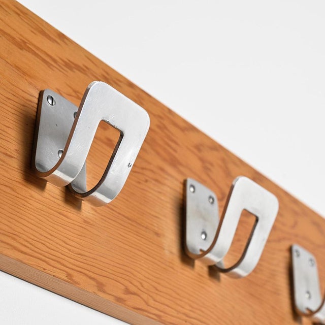 Wall-mounted coat rack composed of a plywood panel veneered in light mahogany, to which four aluminum hooks are attached....