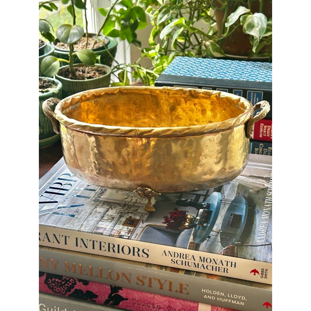 A charming vintage hammered brass cachepot featuring a hand-hammered finish, decorative cast handles, and raised paw feet....