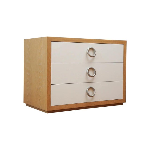 Modern bedside chest of three-drawers shown in light cerused oak frame on plinth base. Fixed round satin nickel pulls on...