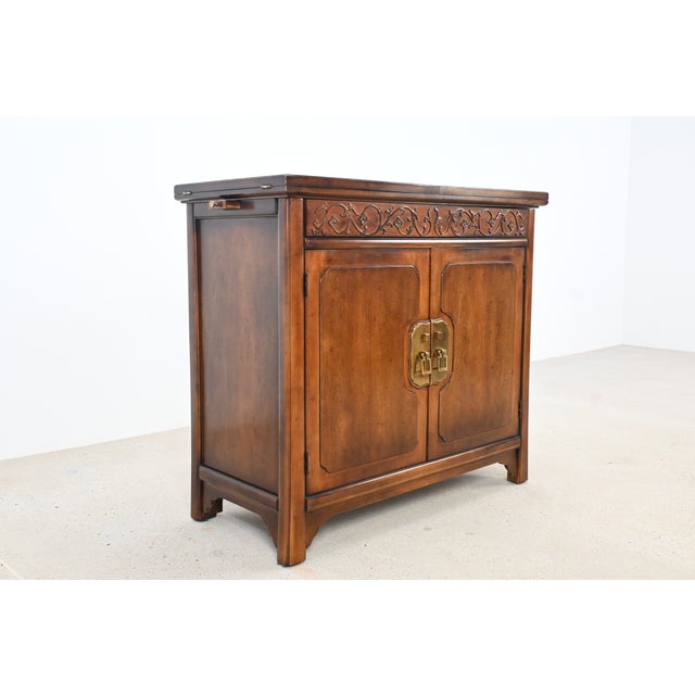 This elegant mid-century Chinoiserie-style drop-leaf bar cabinet features a richly grained wood finish, ornate carved...