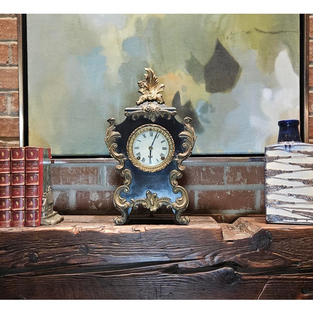 This Ansonia mantle clock, crafted in the rococo style, features a black metal case with cast iron or bronze mounts and...