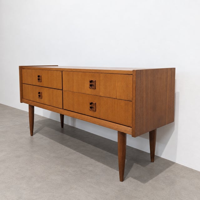 Compact mid-century teak sideboard with four drawers, tapered legs and sculptural organic handles. its balanced...