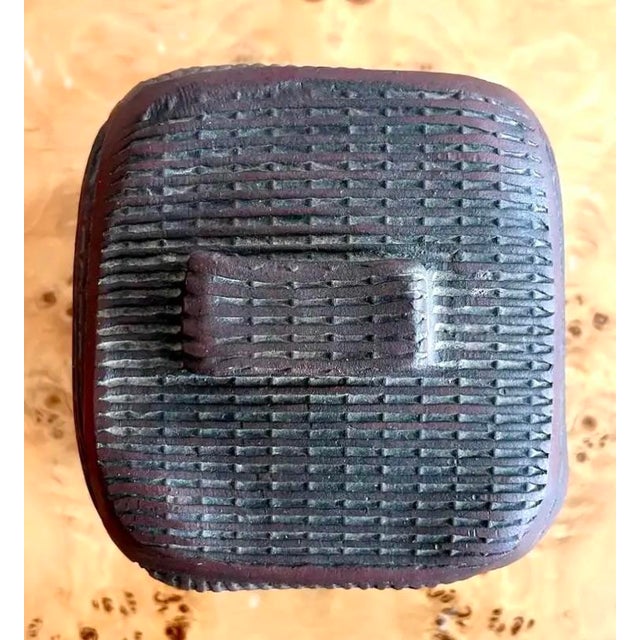 This is a textured ceramic lidded box with a dark, earthy finish and a woven-like surface pattern. Its cube-like form,...