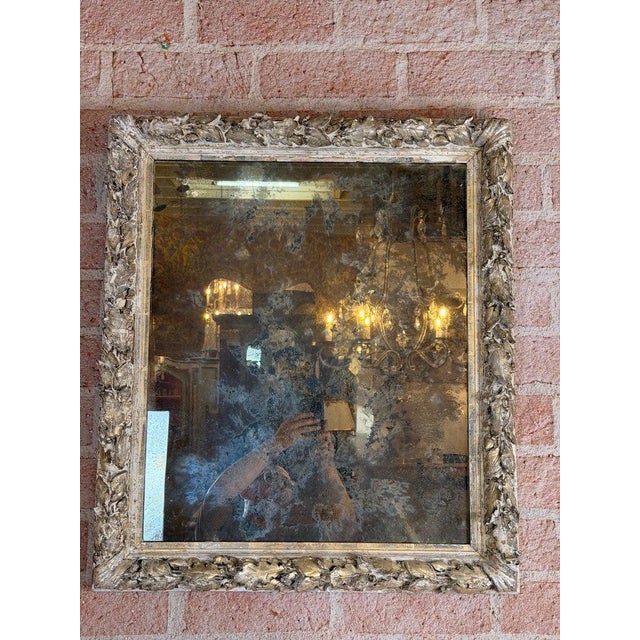 An elegant French Rococo mirror featuring an intricately hand-carved silvered wood frame adorned with flowing foliate and...