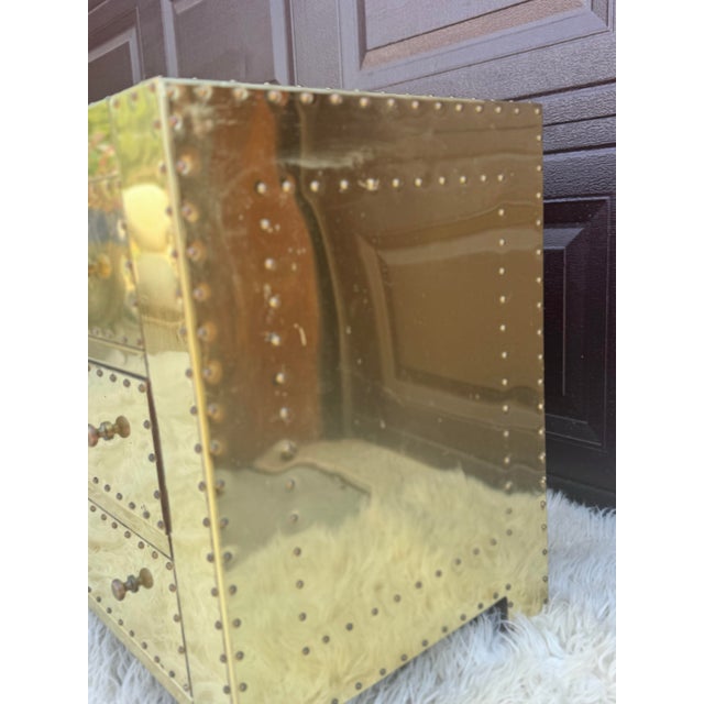A striking pair of vintage Hollywood Regency-style three-drawer nightstands featuring brass-tone metal cladding,...