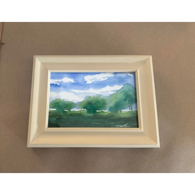 A peaceful original watercolor landscape capturing a sunlit meadow beneath a brilliant blue sky. Soft white clouds drift...