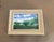 A peaceful original watercolor landscape capturing a sunlit meadow beneath a brilliant blue sky. Soft white clouds drift...