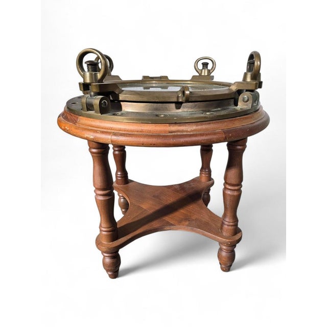This unique side table features a vintage brass ship's porthole repurposed with a glass top, creating a striking nautical-...
