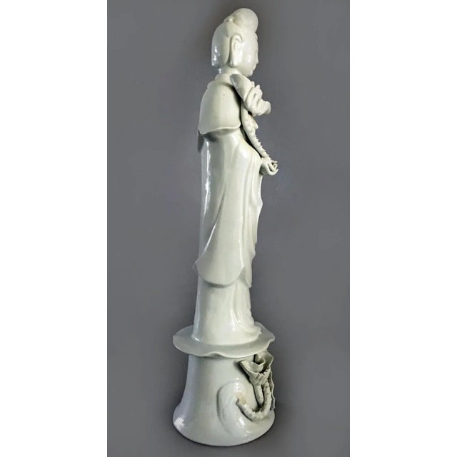 Chinese tall blanc de chine standing Quan Yin dressed in a flowing robe holding a lotus blossom which suggests purity. The...