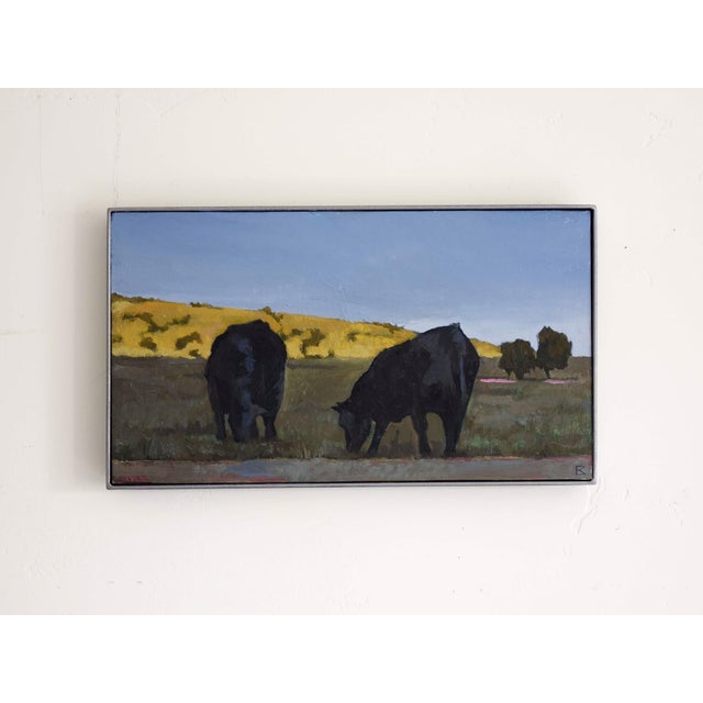 In "Evening Glow," two silhouetted cows graze peacefully in a sunlit field, their dark forms contrasting against the warm,...