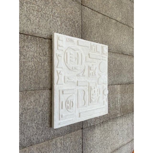 Textured relief wall art with a geometric, grid-like composition. The surface features repeating abstract forms and...